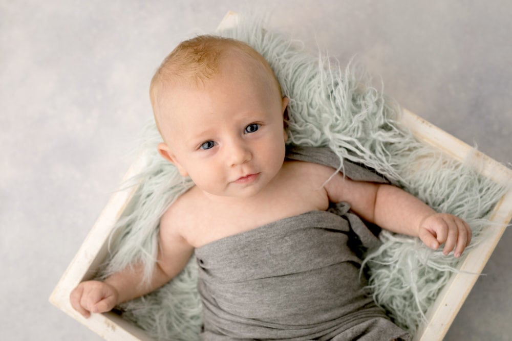 Portrait of a 3 month old baby boy in a grey wrap laying in a white crate