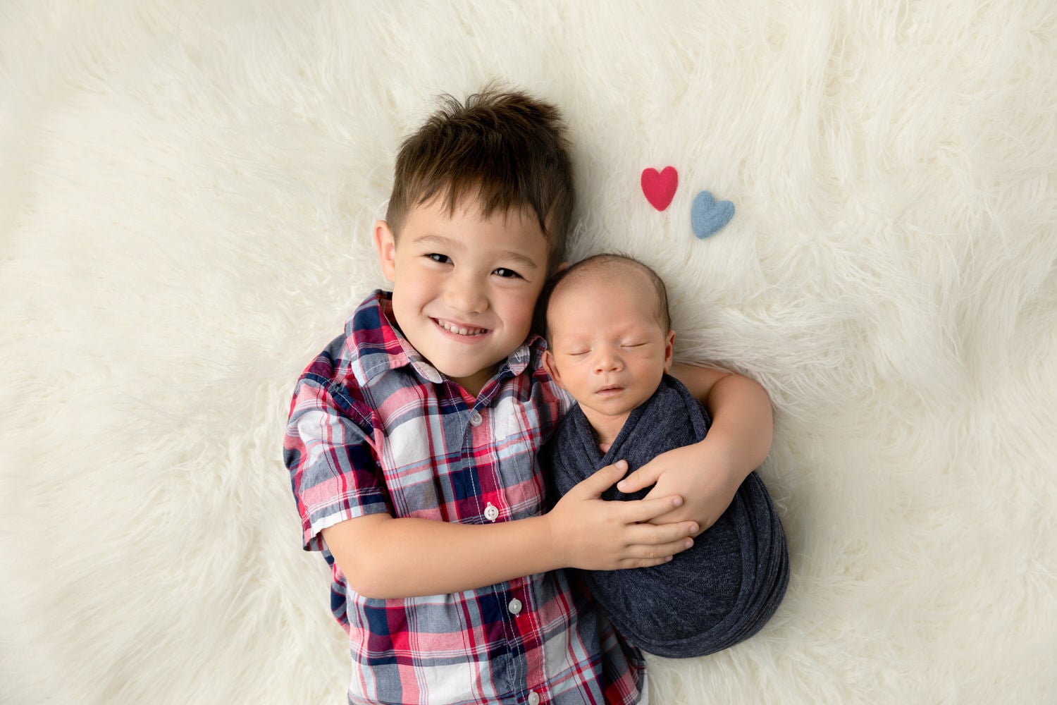 A toddler boy snuggles with his newborn baby brother on a cream colored rug with 2 hearts