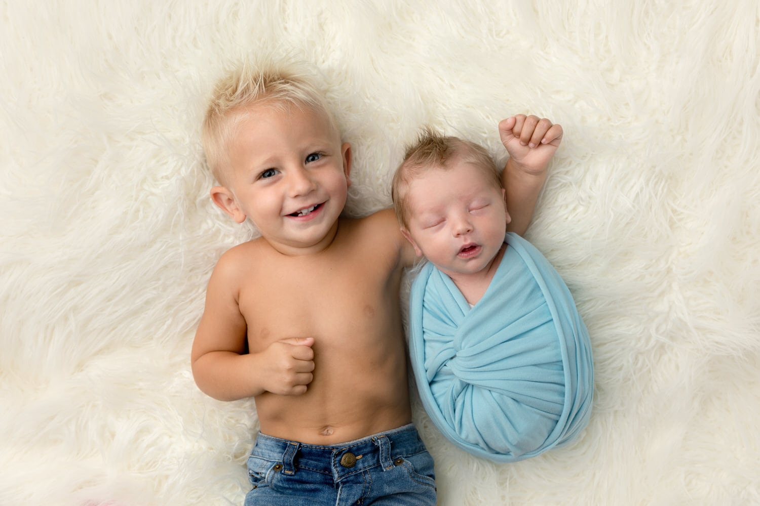 A toddler poses with his newborn baby brother on a cream rug