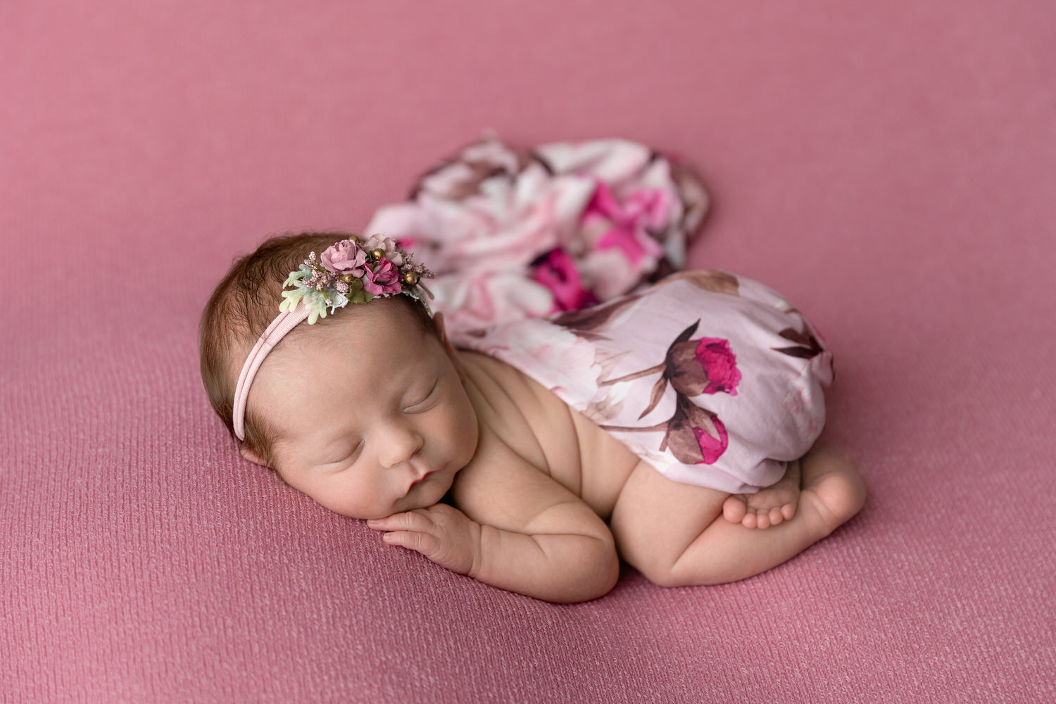 Newborn baby girl sleeps on her belly wrapped in floral fabric on a pink background.
