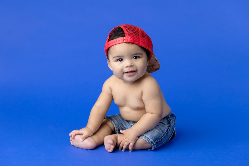6 month baby boy milestone portrait on a blue background with jeans and a red hat