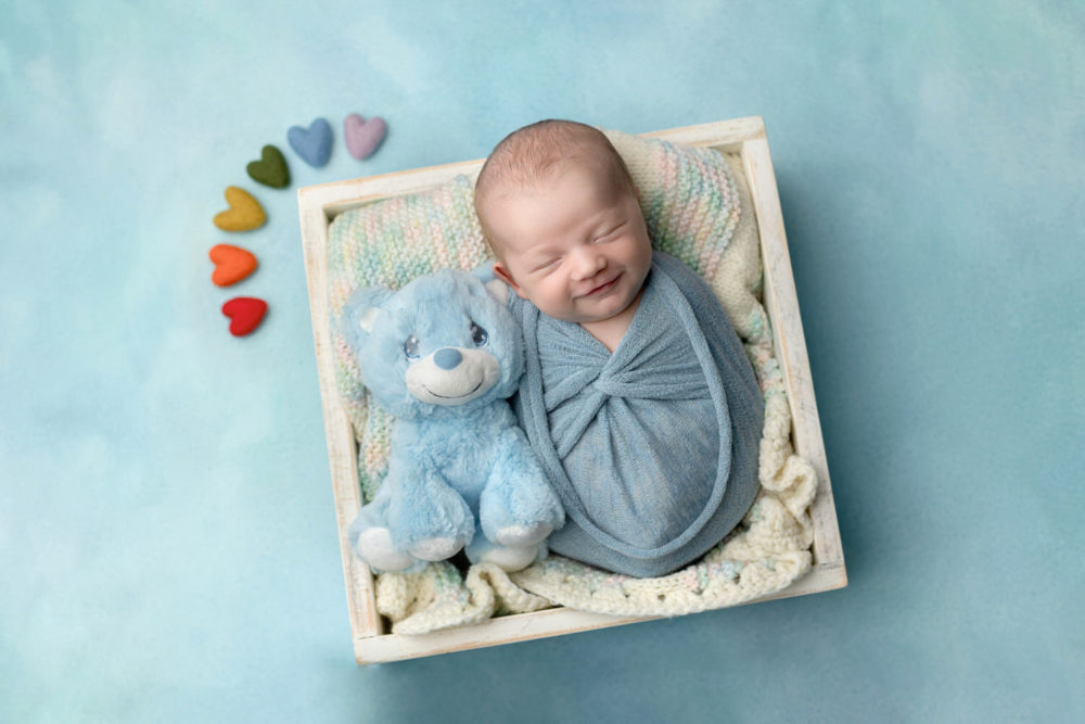 Rainbow newborn baby boy snuggles with his brothers blanket and teddy bear on a light blue background. 