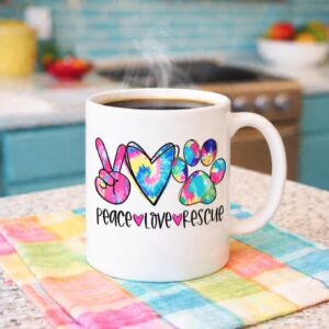 White ceramic coffee mug with colorful Peace Love Rescue design sitting on a vibrant striped towel on a kitchen counter with steam rising from hot coffee